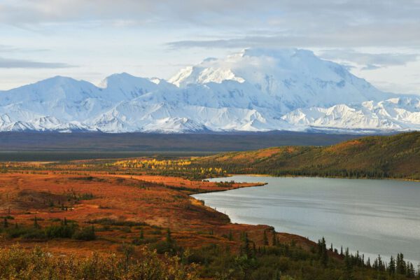 Autumn colors blanket the tundra before a majestic, snow-capped mountain range, with a serene lake nestled in the heart of the valley.