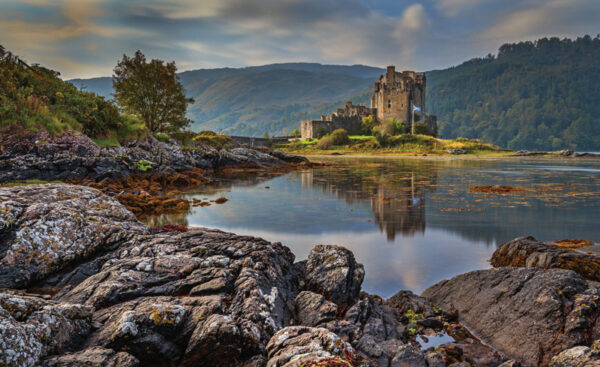 A serene landscape featuring an ancient castle perched on a lush, green peninsula, reflected in the calm waters of a loch, with rocky shores in the foreground and rolling hills shrouded in mist