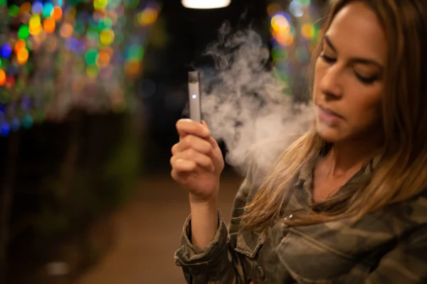 A woman examines a vape pen amidst a haze of exhaled vapor, with colorful bokeh lights in the background, unaware of the risks it poses to venous disease.