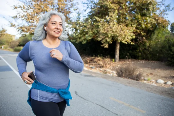 A smiling woman with grey hair enjoying a heart-healthy jog outdoors while holding a smartphone.