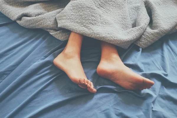 Cozy bare feet protruding from under a soft grey blanket on a blue bedsheet, suggesting a relaxed or lazy day indoors, monitored by the expertise of the Cardiovascular Institute of the South.
