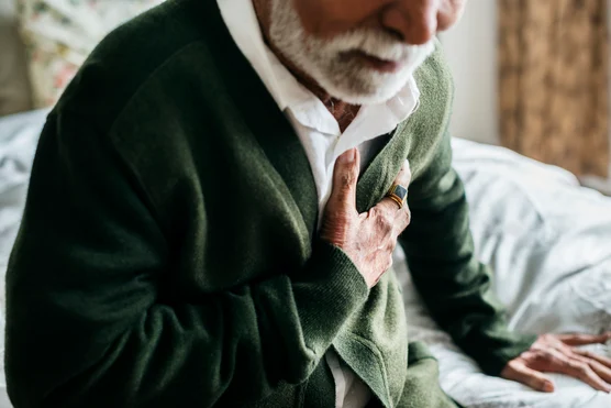 An elderly man in a white shirt and green cardigan thoughtfully resting his hand over his chest, conveying a sense of contemplation about heart health or recollection.