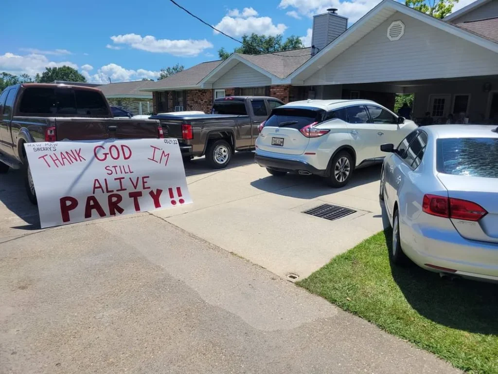 Joyful celebration at a residential driveway with a handmade sign exclaiming 'thank god I'm still alive party!!' hosted by a cardiologist to emphasize heart health.