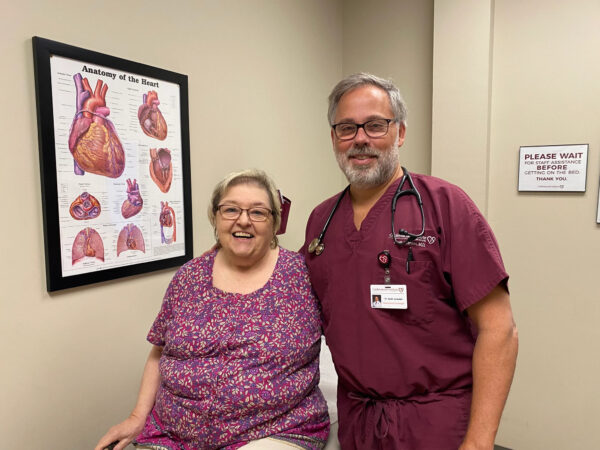 A smiling cardiologist in scrubs stands beside a seated patient, both posing for a photo in a medical office with educational heart anatomy posters on the wall.