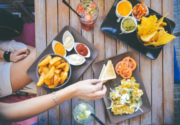 A vibrant array of appetizers on a wooden table, with a person reaching for a piece of bread to complement a heart-healthy meal consisting of wedges, nachos, dips, and a fresh