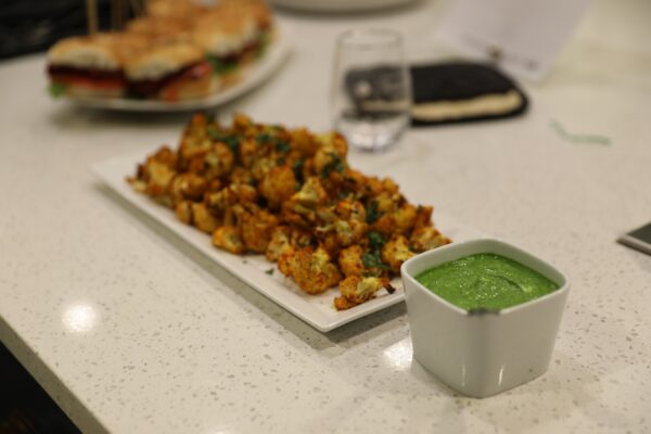 A platter of spiced, roasted cauliflower served with a side of heart-health-focused green sauce, with a blurred background featuring a sandwich and a glass.