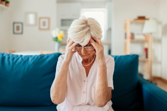 A senior woman sitting on a blue sofa looking stressed or tired as she holds her head in her hands, possibly worried about her venous disease.