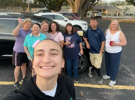 A group of cheerful people posing for a selfie in a parking lot, with several cars in the background and one person playfully waving, celebrating their successful recovery from venous disease.