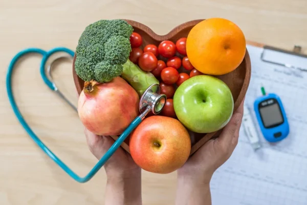 A cardiologist holds a heart-shaped selection of healthy fruits and vegetables with a stethoscope, symbolizing the connection between a nutritious diet and heart health, while a glucometer rests nearby, indicating