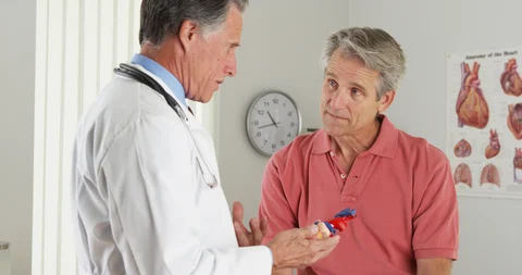 A cardiologist explaining medication to a male patient in a clinic, with a poster of the human heart on the wall in the background.