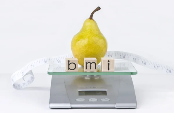 A ripe pear sits atop a digital scale, with the letters "bmi" spelled out in wooden blocks, surrounded by a measuring tape, symbolizing the concept of body mass index and the importance of