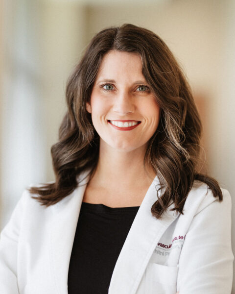 A professional portrait of a smiling woman with long, wavy hair, wearing a white lab coat over business casual attire, possibly representing a heart doctor or scientist at the Cardiovascular Institute of the South.