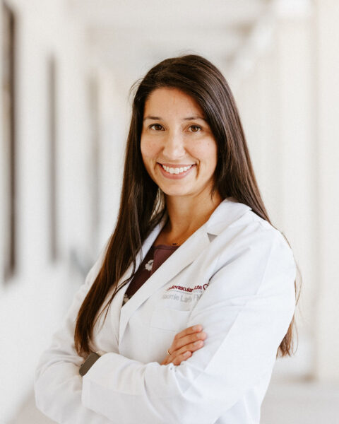A confident heart doctor smiling, with arms crossed, in a white lab coat with a name tag and credentials.