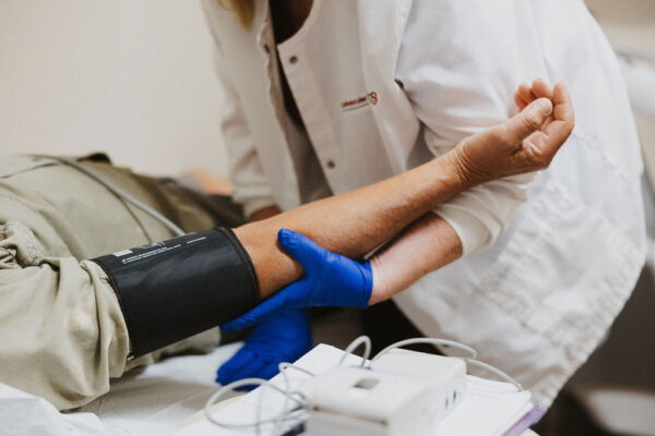 A cardiology health professional in a white coat and blue gloves is taking a patient's blood pressure using a sphygmomanometer.