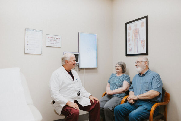 A cardiologist, seated, engaging in a friendly conversation with an elderly couple in a medical consultation room.