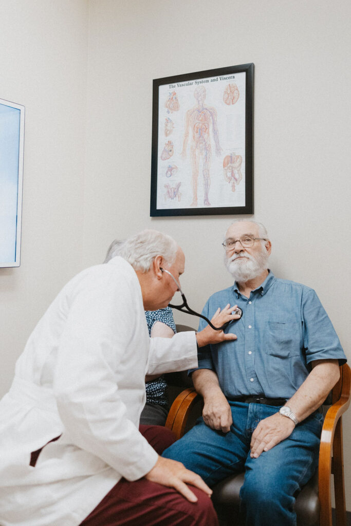 A heart doctor in a white coat using a stethoscope to listen to the chest of a male patient seated in the medical office of the Cardiovascular Institute of the South, with an anatomical poster