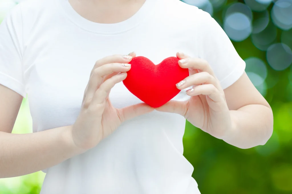 A person holding a red heart-shaped object with both hands, symbolizing heart health and love, against a blurred natural green backdrop.