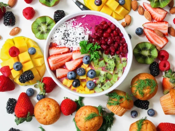 A vibrant and colorful bowl of fruit smoothie garnished with a variety of fresh fruits, nuts, and coconut, accompanied by fluffy muffins on a bright white background, perfect for heart health.