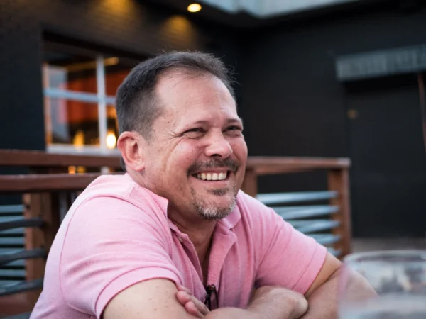 A man in a pink shirt enjoying a hearty laugh while sitting outside at a casual dining area during evening time promotes heart health.