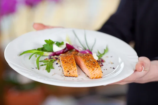 Waiter presenting a heart-healthy gourmet dish of grilled salmon with fresh herbs and garnishes on a white plate.
