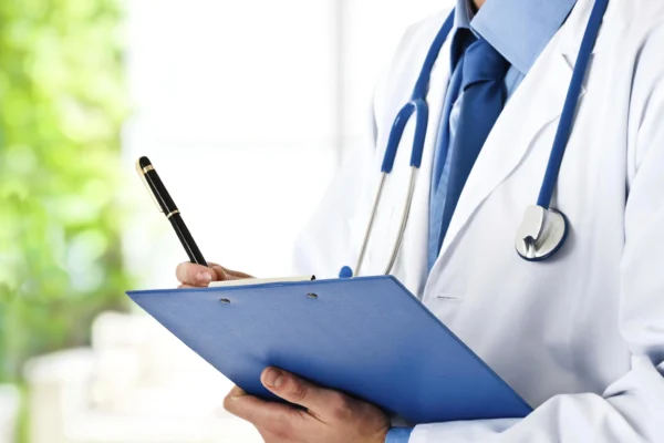 A cardiologist in a white coat with a stethoscope stands holding a clipboard and a pen, ready to review heart health records or take patient notes.