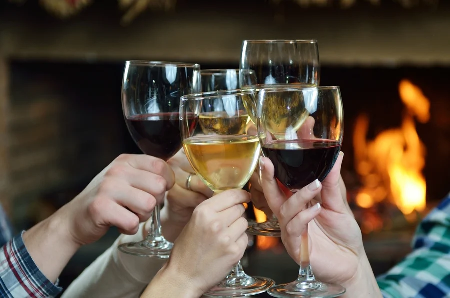 Cheers to warmth and good company: friends sharing a toast with glasses of red and white wine by a cozy fireplace, celebrating the benefits of moderate wine consumption as noted by cardiology research.