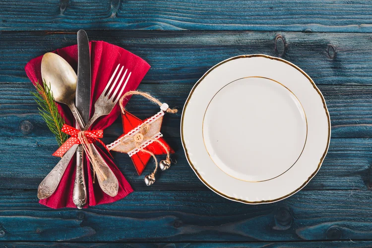 A festive table setting with vintage silverware tied with a red ribbon and a sprig of evergreen on a red napkin, next to an elegant plate with a gold rim on a dark blue wooden