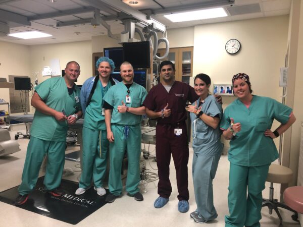 A group of happy looking hospital staff in scrubs gives the thumbs up in an operating room