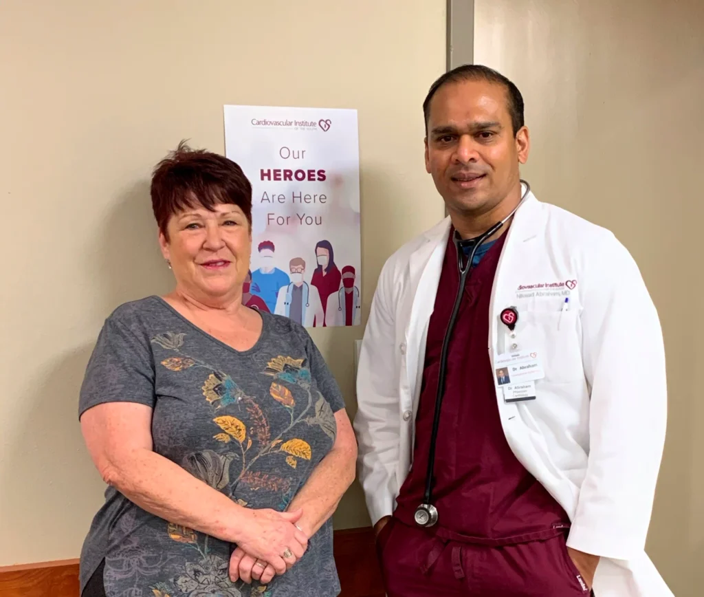 A smiling healthcare professional standing beside a patient, in front of a sign that reads "our heroes are here for you" at the Cardiovascular Institute of the South.