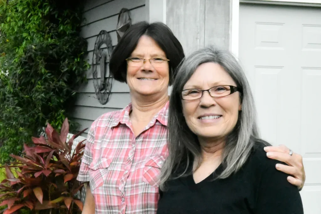 Two smiling women standing together in a friendly embrace outside a house with greenery and a decorative wheel on the wall in the background, symbolizing heart health.