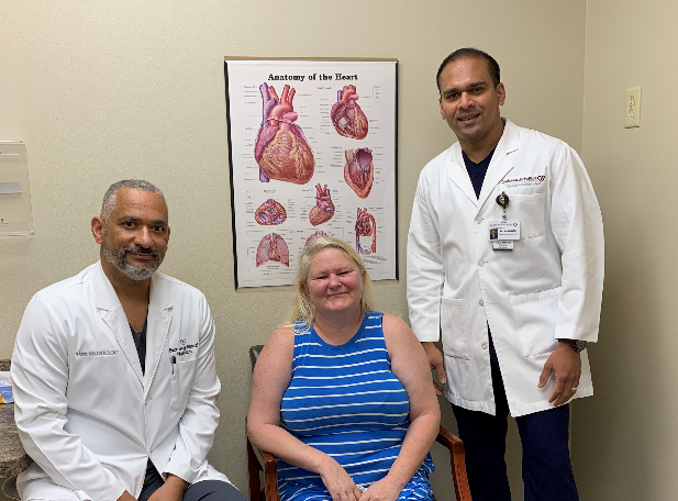 Two healthcare professionals in white coats posing with a patient in a cardiology clinic, with an educational poster of the heart's anatomy in the background.