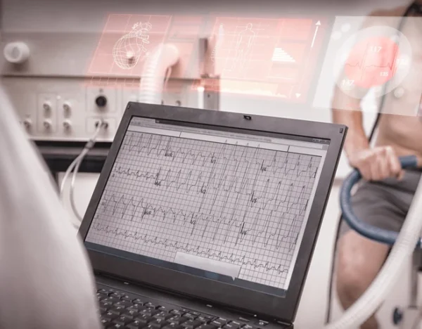 A cardiologist studying a laptop displaying an electrocardiogram (ECG) readout in a clinical setting, with a patient undergoing a cardiac test in the background.