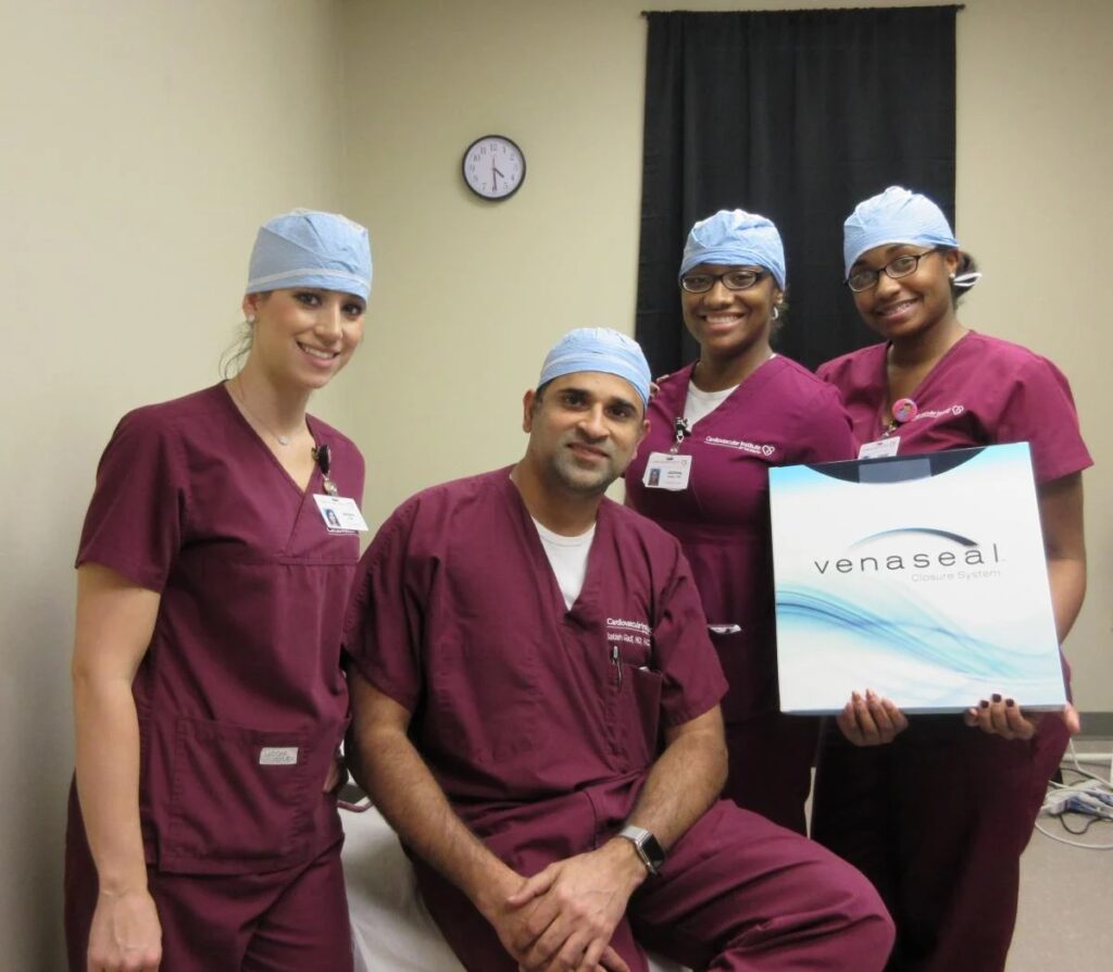 A team of healthcare professionals, including a cardiologist in burgundy scrubs and surgical caps, posing for a photo in a clinical setting, with one member seated and holding a promotional board for a medical