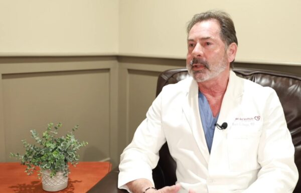 A professional, likely a heart doctor, in a white lab coat sits at a table during an interview or discussion, with a small potted plant on the table adding a touch of greenery to the