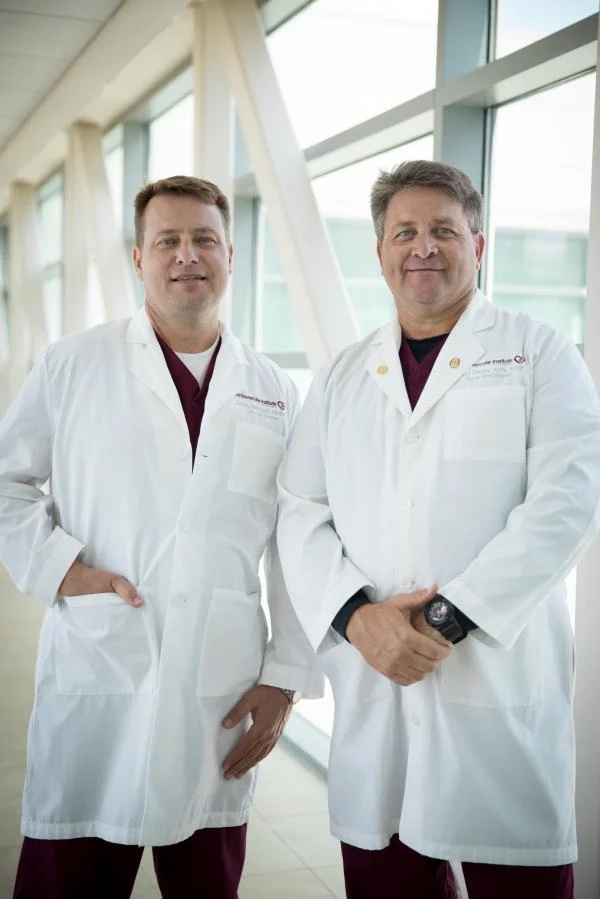 Two cardiologists standing confidently in a bright corridor of the Cardiovascular Institute of the South, wearing white lab coats over maroon scrubs.