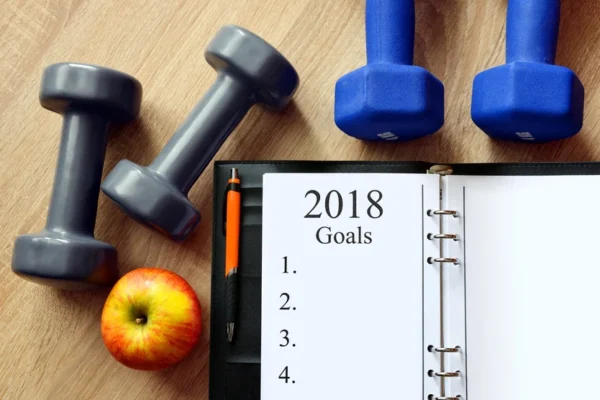 Fitness planning concept with dumbbells, an apple, and a list of goals for 2018 recommended by a cardiologist on a notebook.