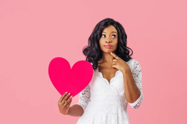 A woman in a white dress holding a red, heart-shaped cutout symbolizing cardiology while looking pensive against a pink background.
