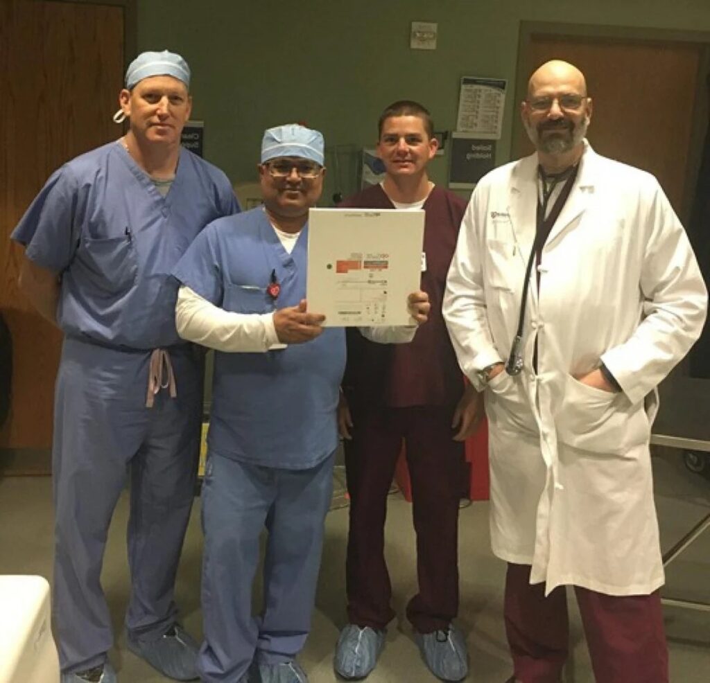 Four cardiology professionals posing for a photo in a hospital setting, with one of them holding up a certificate.