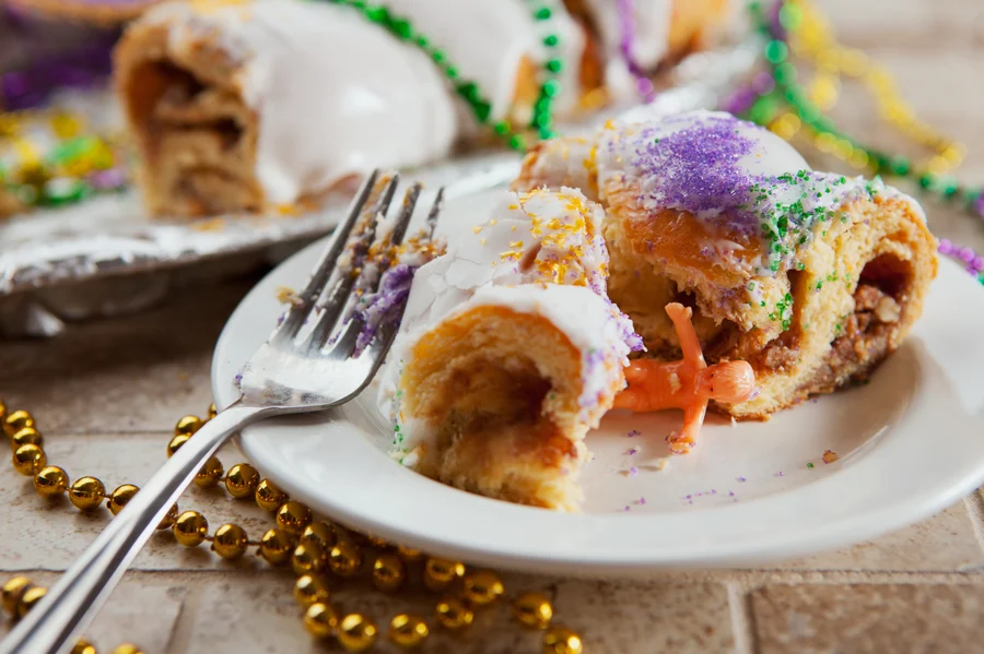 A delicious slice of traditional mardi gras king cake with a hidden baby figurine on a plate, complete with festive beads and endorsed by the Cardiovascular Institute of the South, ready for the next bite