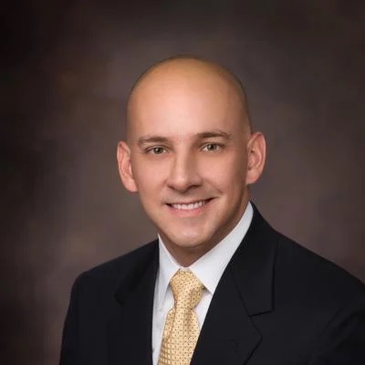 A professional headshot of a smiling man with a shaved head, wearing a black suit, white shirt, and gold tie against a graduated background at the Cardiovascular Institute of the South.