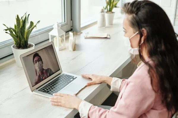 A woman in a face mask engaged in a video call on her laptop with a cardiologist who appears on the screen, exemplifying remote communication in health-conscious times.