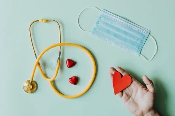 A healthcare-themed composition featuring a yellow stethoscope, two red heart-shaped objects, a surgical face mask, and a hand holding a paper heart against a soft green background, symbolizing care and medical