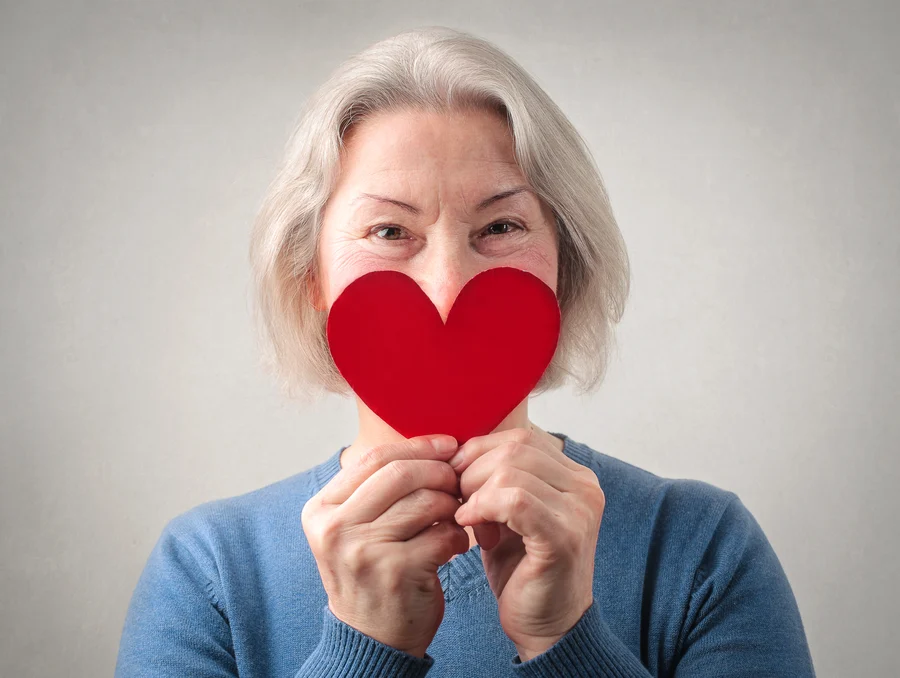 Elderly woman smiling gently behind a red heart-shaped card, symbolizing love, care, or affection for cardiovascular health.