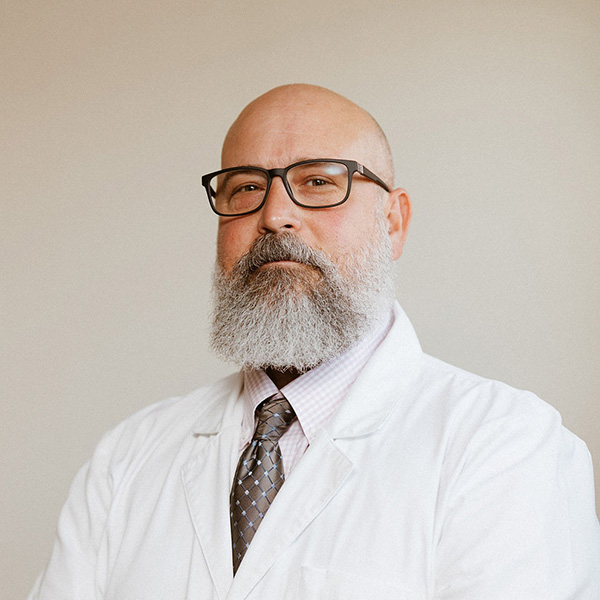 A poised individual wearing glasses with a full beard, dressed in a white lab coat over a shirt and tie, conveying an air of experience and professionalism.