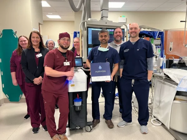 A team of smiling medical professionals standing in an operating room with surgical equipment, including one holding a certificate next to the aurion machine.
