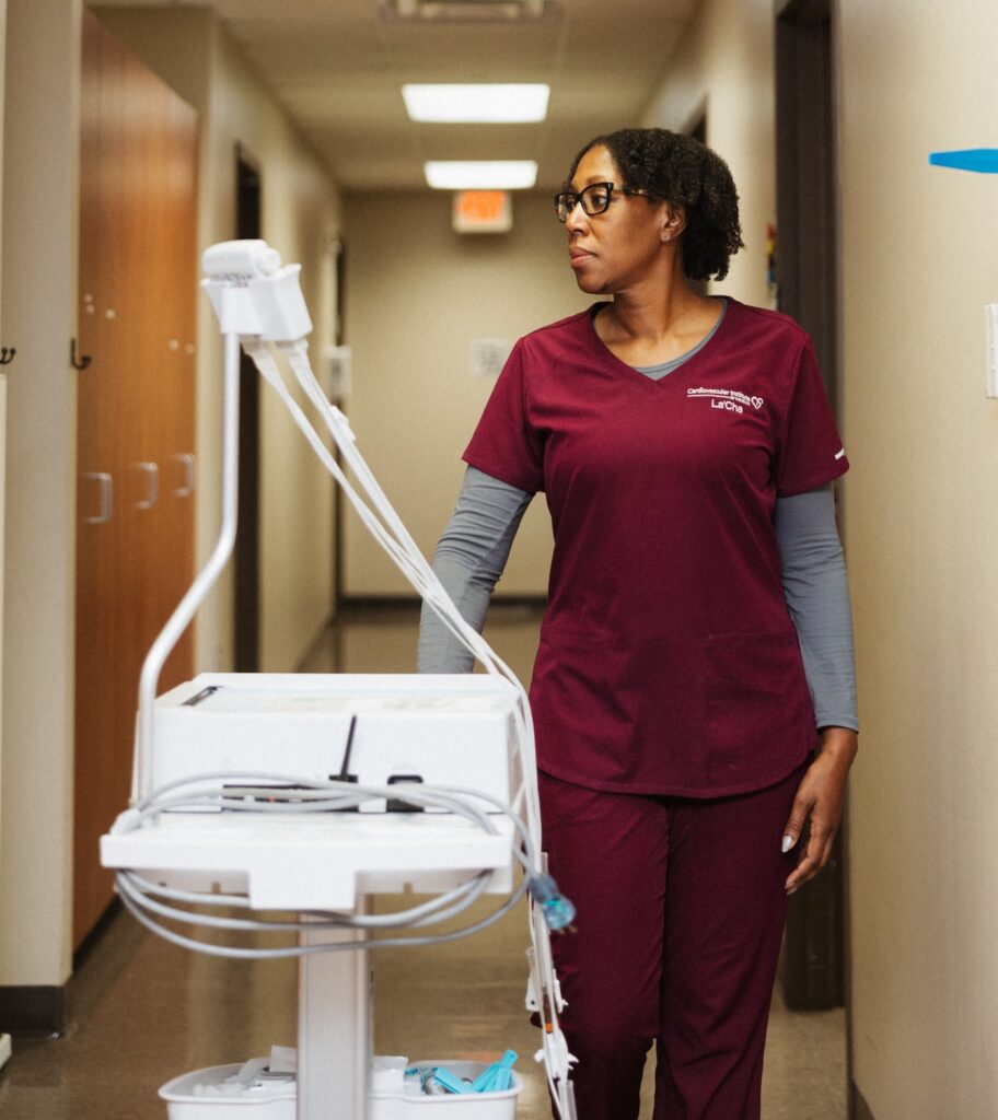 Healthcare worker in burgundy scrubs stands in a hallway next to medical equipment on a cart, looking to the side.