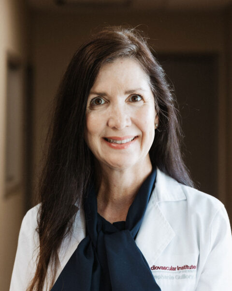 A smiling woman with long brown hair wearing a white lab coat over a navy blouse, identified as a professional at a cardiovascular institute.