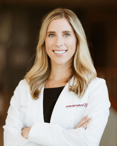 Professional portrait of a smiling woman with blonde hair, wearing a white lab coat with a badge that reads "cardiovascular institute." she stands with her arms crossed.