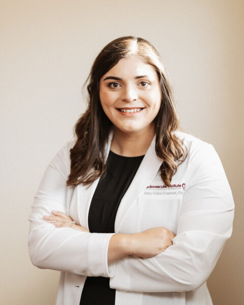 Confident professional woman standing against a neutral background with crossed arms, wearing a white lab coat indicating a medical or scientific profession.