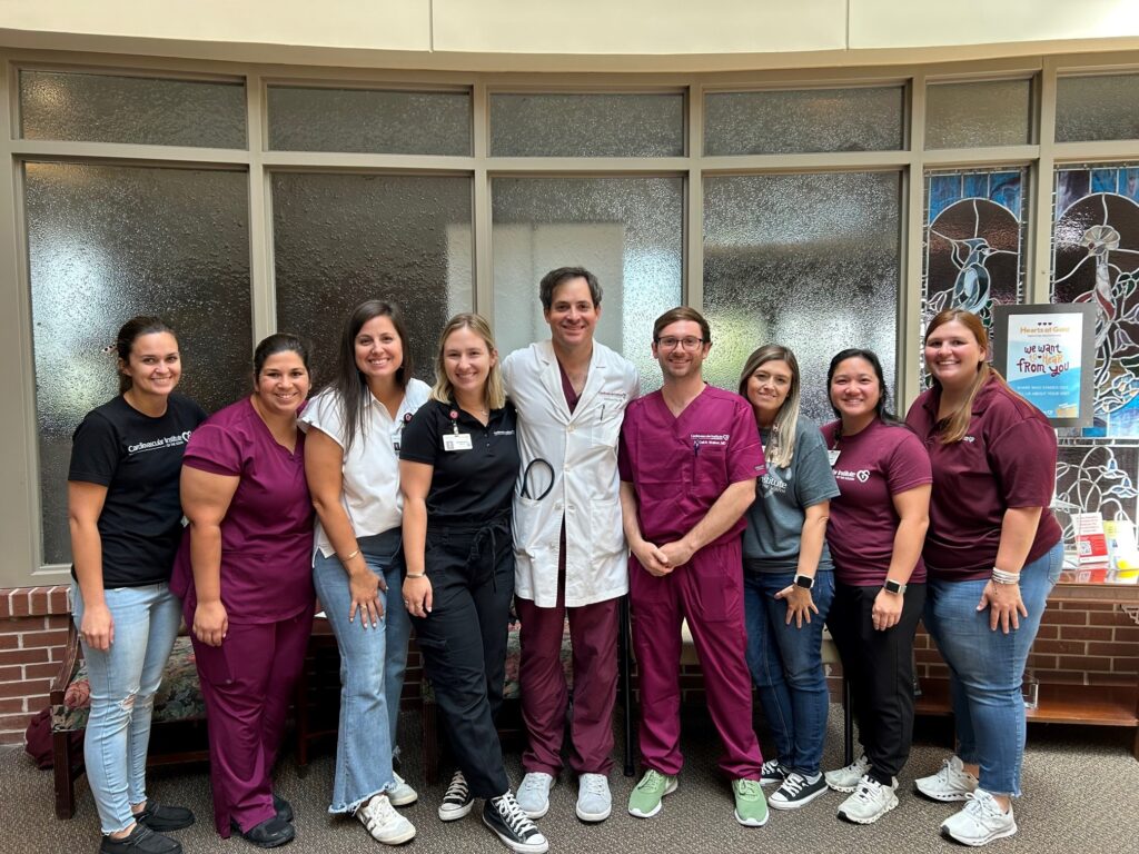A group of nine medical professionals, wearing a mix of scrubs and casual attire, poses for a photo with smiles, showcasing a sense of teamwork and camaraderie in a bright, indoor setting with frosted glass windows in the background.